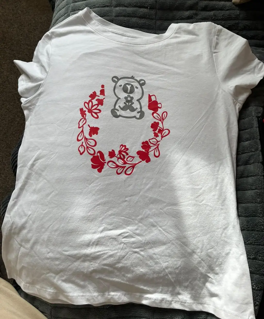 A white t-shirt printed with a grey cuddly animal holding a flower inside a red floral wreath
