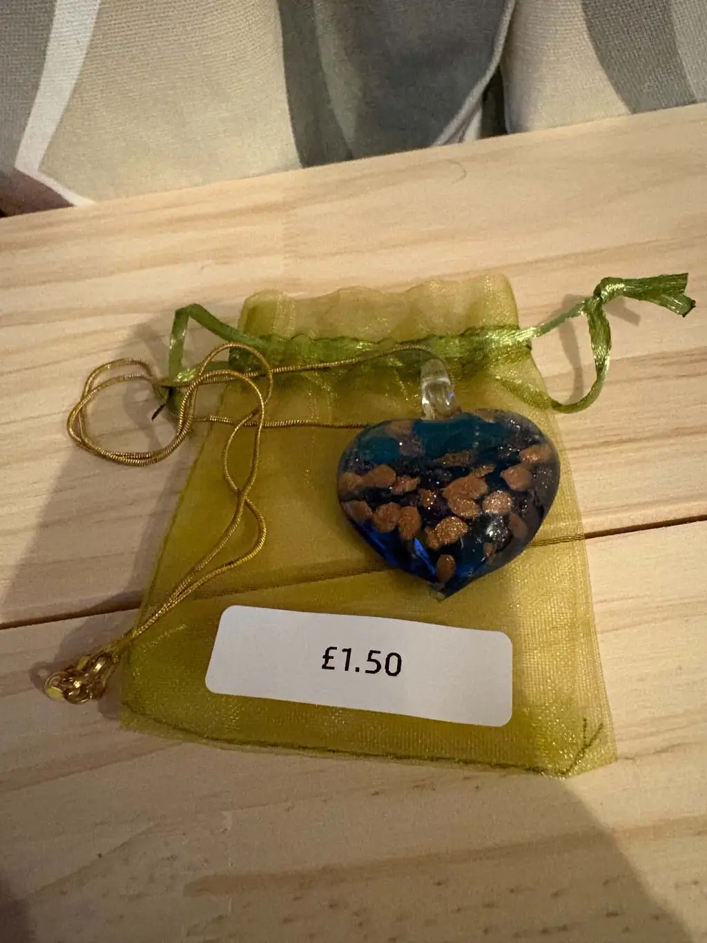 A blue glass heart pendant flecked with gold on a gold chain, on a green organza bag