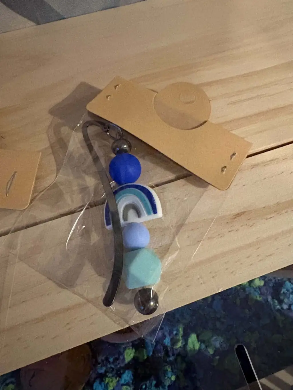 A metal bookmark with a blue rainbow charm and blue silicone beads, in a clear bag