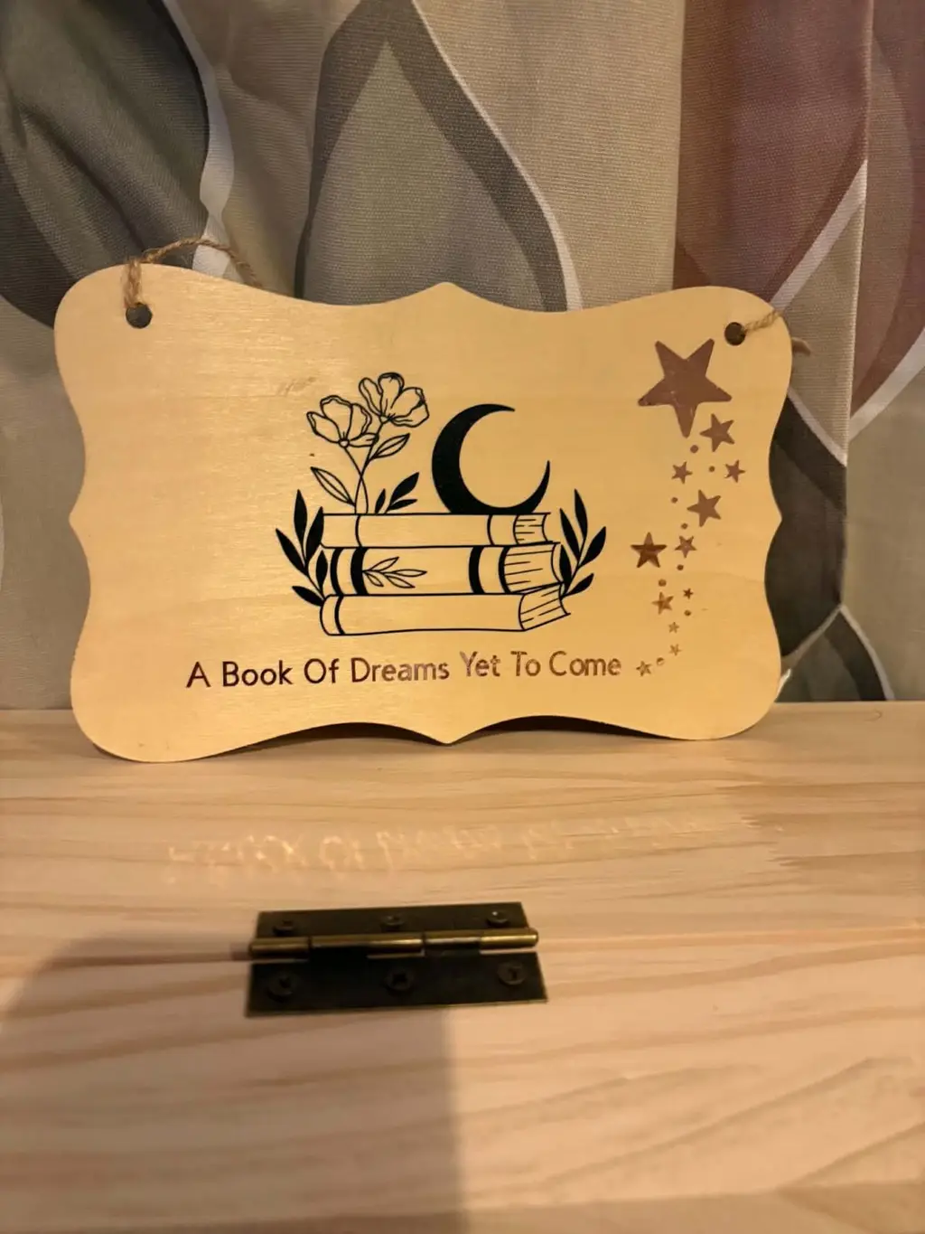 A wooden hanging plaque with a stack of books, flowers, a moon and stars, and the words A Book Of Dreams Yet To Come
